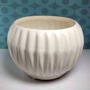 May include: A round, off-white ceramic planter featuring a series of vertical, recessed fluted grooves around its exterior. The bowl-shaped vessel has a smooth, glossy finish and a wide, open top, providing a clean and simple aesthetic for plants.