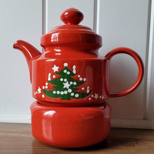May include: A bright orange ceramic teapot with a matching lid and a separate circular base. The teapot features a green Christmas tree graphic decorated with white dots and stars. The design includes a curved handle and a short spout.