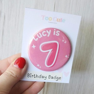 Puede incluir: Insignia de cumpleaños rosa con el texto "Lucy is 7" en letras blancas. El número 7 está en una fuente rosa, como de burbuja. La insignia tiene un efecto brillante.