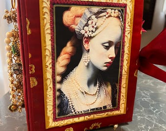 May include: A red velvet-covered book with a framed portrait of a woman in a historical dress. The portrait is surrounded by a gold frame. The book has gold embellishments and a decorative charm. A red ribbon is attached to the top.