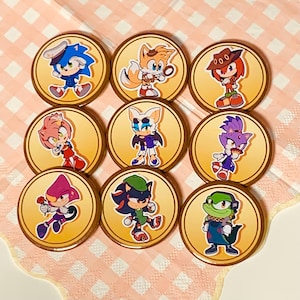 May include: Nine round pins featuring different characters from the Sonic the Hedgehog franchise. Each pin depicts a different character in a colourful cartoon style against a yellow background. The characters include Sonic, Tails, Knuckles, Amy, Shadow, Rouge, Blaze, Vector, and Espio.