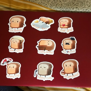 May include: Nine stickers featuring cartoon bread characters with different expressions and puns. The stickers are on a red laptop. The stickers include "I'm Bready", "bread time", "I loaf you!", "Just a glaze", "Loafing Around", "Le pain", "Grainy Day", "Feeling Crusty", and "All jam packed!"