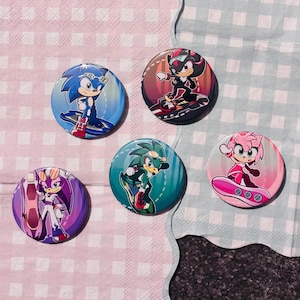 May include: A collection of five round buttons featuring characters from the Sonic the Hedgehog video game series. Each button has a different character illustration, with vibrant colors and dynamic poses. The buttons are set against a pink and blue checkered background.