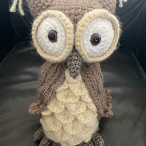 以下が含まれることがあります： 茶色とベージュの糸で編まれた、フクロウのぬいぐるみ。フクロウは、茶色の瞳がついた大きな白い目と、茶色のくちばしをしています。