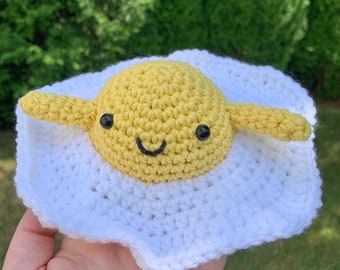 Puede incluir: Un juguete de peluche tejido a crochet con forma de huevo frito. La yema es amarilla con una cara sonriente y ojos negros. La parte blanca del huevo está tejida a crochet con hilo blanco.