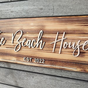 Op de afbeelding: Een rustiek houten bord met de woorden "The Beach House" in witte cursieve letters en "EST 2022" eronder. Het hout heeft een gebrande afwerking.