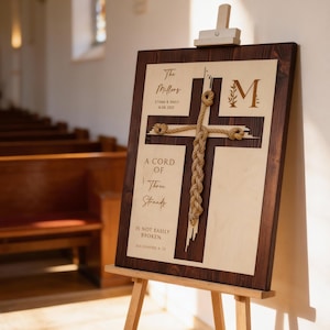 May include: A wooden sign with a cross design, featuring rope accents and the text "A Cord of Three Strands Is Not Easily Broken." The sign is displayed on a wooden easel, with the names "The Millers" and a date visible.