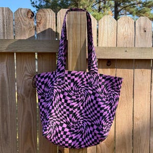 Puede incluir: Un bolso tote con un patrón de cuadros negros y rosas. El bolso tiene un diseño en espiral y dos asas. El bolso está colgado de una valla de madera.