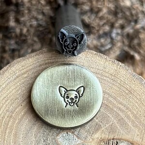 May include: A metal stamp with a chihuahua face design. The stamp is shown with an impression on a round metal disc. The stamp is dark gray, and the disc is a golden color. The background is a wooden surface.