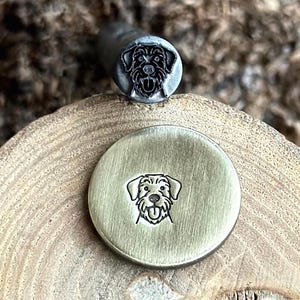 May include: Two metal stamps featuring dog designs. One is a round stamp with a dog face engraving. The other is a cylindrical handle with a dog face engraved on the end. The stamps are on a wooden surface.