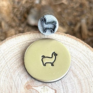 Könnte beinhalten: Ein Metallstempel und eine gestempelte Messingscheibe, beide mit einem Lama-Design. Der Stempel ist grau und die Scheibe goldfarben. Das Lama-Bild ist eine einfache Umrisszeichnung. Die Artikel werden auf einer Holzoberfläche präsentiert.