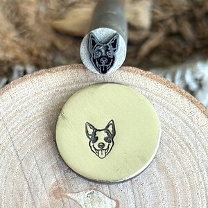May include: A metal stamp with a black and white image of a dog's head. The stamp is on a wooden surface and has been used to create an impression of the dog's head on a round piece of metal.