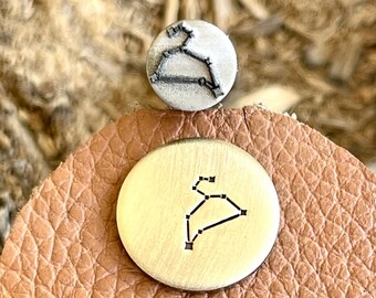 May include: A gold-toned metal charm with a constellation design. The charm is on a brown leather background.