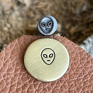 May include: A close-up of a small, round, brass stamp with an alien face design. The stamp is on a piece of brown leather.