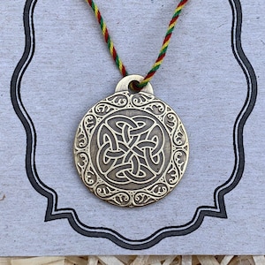 Può includere: Un ciondolo in ottone con un designo a nodo celtico, appeso a un cordoncino verde, rosso e giallo. Il ciondolo è rotondo e presenta un design dettagliato e intricato.