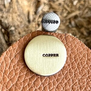 May include: A handcrafted leather earring with a copper disc and a small silver ball. The copper disc is stamped with the word "COPPER". The silver ball is stamped with the word "essence". The earring has a textured, brown leather backing.