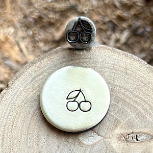 Puede incluir: Un sello de metal con forma de cereza y un círculo estampado con un diseño de cereza. El sello es plateado y el círculo estampado es dorado. El diseño de la cereza es un simple contorno de dos cerezas y una hoja.