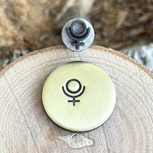 May include: A small, silver metal stamp and a round, light yellow metal disc featuring the astrological symbol for Pluto. The stamp is above the disc on a wooden surface, with a blurred background.