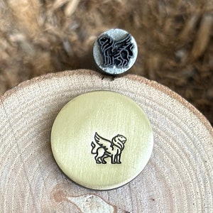 May include: A brass stamp with a winged lion design. The stamp is on a wooden surface.