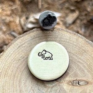 Puede incluir: Un primer plano de un sello de metal con un diseño de mamut. El sello está sobre una superficie de madera y se ha utilizado para crear una impresión del mamut en un pequeño disco redondo de color crema.