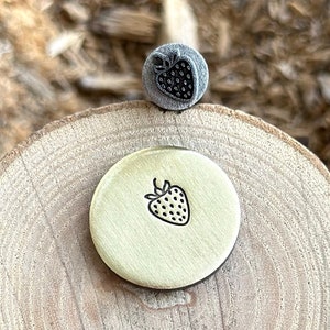 Op de afbeelding: Een close-up van een messing stempel met een aardbeidesontwerp. De stempel ligt op een houten oppervlak.