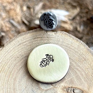 Könnte beinhalten: Nahaufnahme von zwei Metallstempeln, einer mit einem Ameisenmotiv und der andere mit einem Ameisenmotiv. Die Stempel befinden sich auf einer Holzoberfläche.