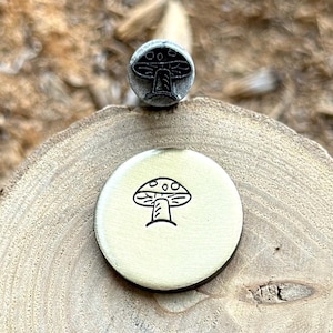 Puede incluir: Un sello de metal con forma de hongo y un disco de latón estampado. El sello es de color plateado y presenta un diseño de hongo. El disco tiene una ilustración de hongo en tinta negra. Los artículos se exhiben sobre una superficie de madera.