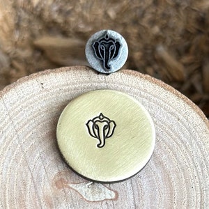 May include: A metal stamp and a stamped brass disc, both featuring a detailed elephant head design. The stamp is gray, while the disc is gold-colored. The objects are placed on a wooden surface, showcasing the stamp's impression.