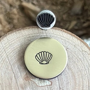 May include: A brass stamp with a seashell design. The stamp is on a wooden surface.
