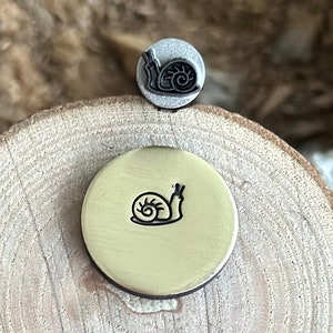 May include: Two metal stamps, one small and one larger, each featuring a snail design. The stamps are positioned on a wooden surface, with the larger stamp resting on a round, brass-colored base. The snail design is in black.