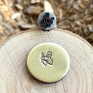 Könnte beinhalten: Ein runder, messingfarbener Metallstempel mit detailliertem Bienenmuster und ein kleinerer, grauer Stempel mit einem Bienenmuster. Die Stempel sind auf einer Holzoberfläche mit sichtbarer Maserung und Textur angeordnet. Das Bienenmuster ist ein häufiges Thema.
