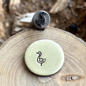 May include: A metal stamp with a goose design. The stamp is on a wooden surface and has been used to create an impression of the goose on a metal disc.