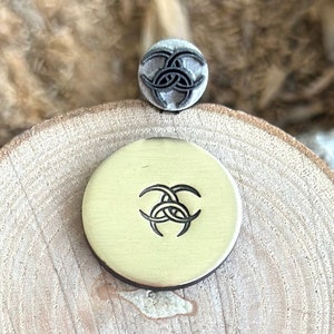 May include: A metal stamp and a brass disc featuring a Celtic knot design. The stamp is small and grey, while the disc is round and gold-coloured. The design is a series of intertwined shapes.