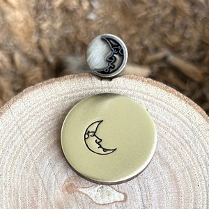 May include: Two celestial-themed metal objects rest on a wooden surface. One is a small, silver-toned button with a crescent moon design. The other is a larger, gold-toned disc with a similar moon face. The objects are likely jewelry or craft supplies.