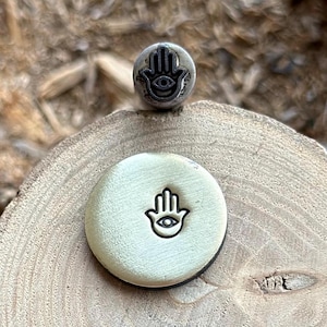 Op de afbeelding: Een close-up van een messing stempel met een hamsa-handontwerp met een oog in het midden. De stempel ligt op een houten oppervlak.