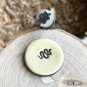 May include: A close-up of a brass stamp with a snake design. The stamp is sitting on a wooden surface.