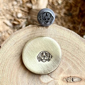 May include: A close-up of a metal stamp featuring a dog's head design. The stamp is resting on a wooden surface. This stamp is used to create bespoke designs on leather or metal.