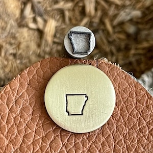 Może przedstawiać: Metalowa pieczątka i mosiężny krążek z konturem stanu Arkansas. Pieczątka jest mała i okrągła, ze szczegółowym obrysem stanu. Krążek jest przymocowany do kawałka brązowej skóry.