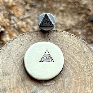 Può includere: Un timbro in metallo argentato con un disegno di occhio onniveggente nero all'interno di un triangolo. Il timbro è su una superficie di legno.