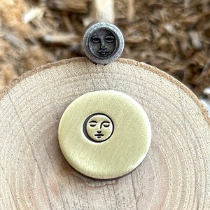 May include: Two metal stamps with a face design. The stamp on the left is gold and the stamp on the right is silver.