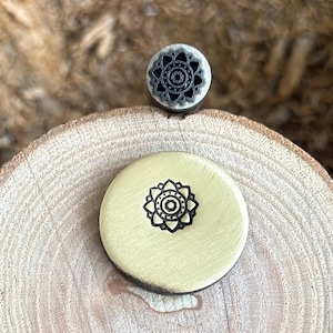 May include: Two round wooden stamps with mandala designs. The top stamp is silver with a black mandala design. The bottom stamp is gold with a black mandala design.