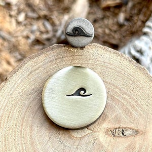 Puede incluir: Dos sellos metálicos con un diseño de onda. El sello más grande es de color dorado y el más pequeño es de color plateado.