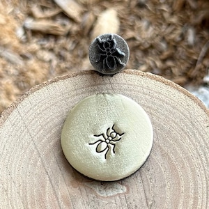 Puede incluir: Un primer plano de un sello de metal con un diseño de hormiga. El sello está sobre una superficie de madera y se ha utilizado para crear una impresión de una hormiga en un pequeño trozo de metal redondo.