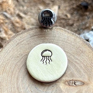 May include: A close-up of a metal stamp with a jellyfish design. The stamp is on a wooden surface, and there is a smaller, stamped jellyfish image on a metal disc.