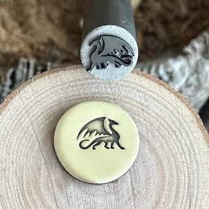 Op de afbeelding: Een close-up van een metalen stempel met een drakenontwerp. De stempel bevindt zich op een houten oppervlak en is gebruikt om een ​​indruk van de draak op een metalen schijf te maken.