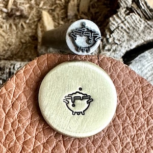 May include: A close-up of a metal stamp with a design of a cauldron with bubbles. The stamp is on a brown leather surface.