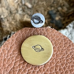 Peut inclure: Gros plan d'un petit tampon rond en laiton avec un motif de planète avec des anneaux. Le tampon est sur un morceau de cuir brun.