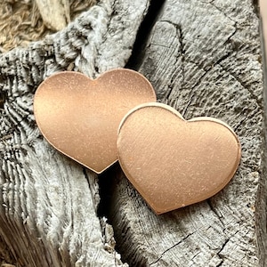 Puede incluir: Dos amuletos en forma de corazón de oro rosa sobre un fondo de madera rústico.
