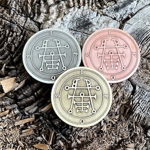 May include: Three round metal tokens with a symbol and letters on them. The tokens are silver, copper, and gold in color. The tokens are on a wooden surface.