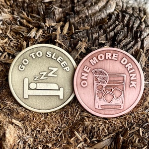 Peut inclure: Deux jetons ronds en métal. L'un est doré avec le texte "Go to Sleep" et une personne dormant dans un lit. L'autre est en cuivre avec le texte "One More Drink" et un verre de glace avec un quartier de citron vert.